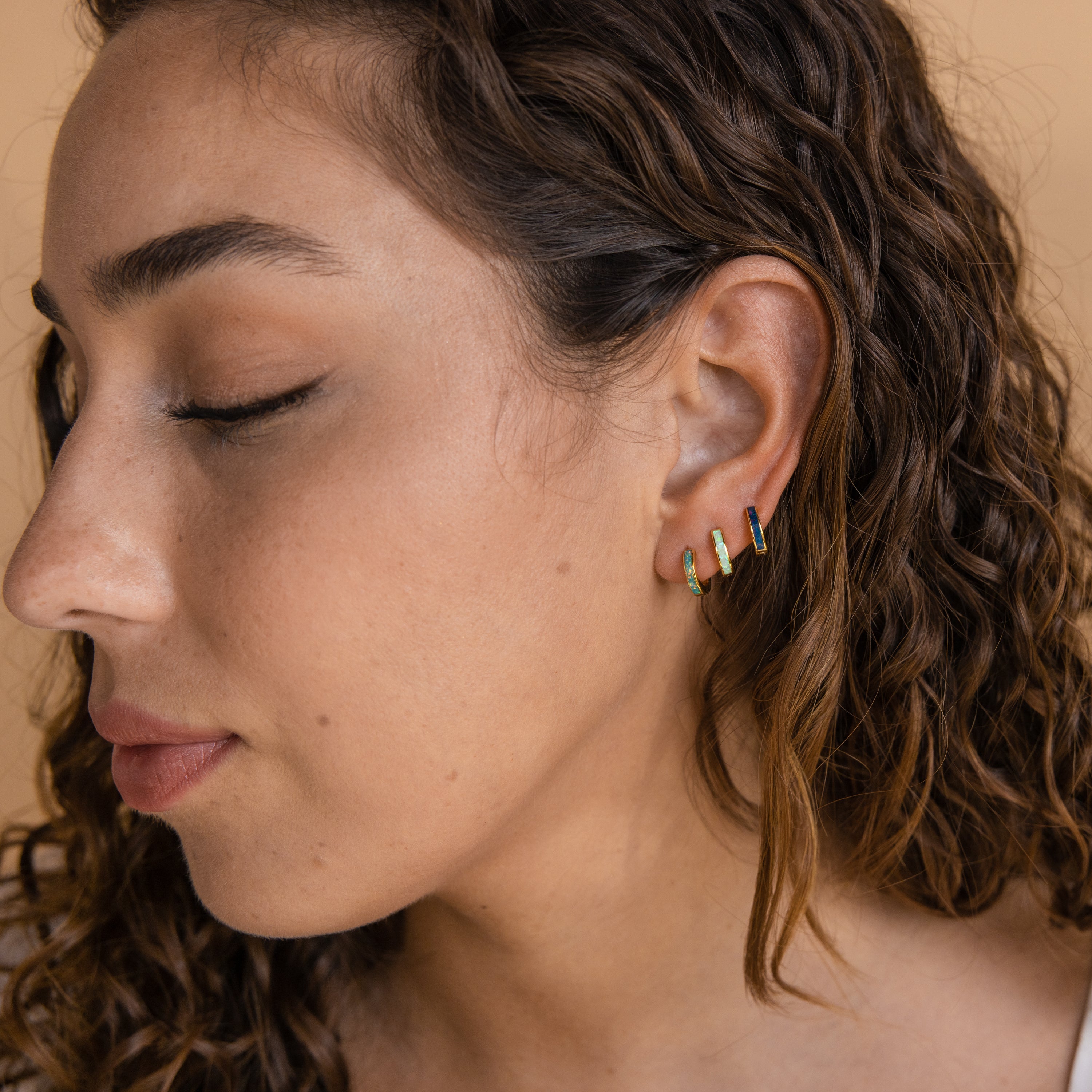 A woman with curly hair and closed eyes wears the Opal Inlay Huggies Set on her left ear, each hoop shimmering with subtle opal colors.