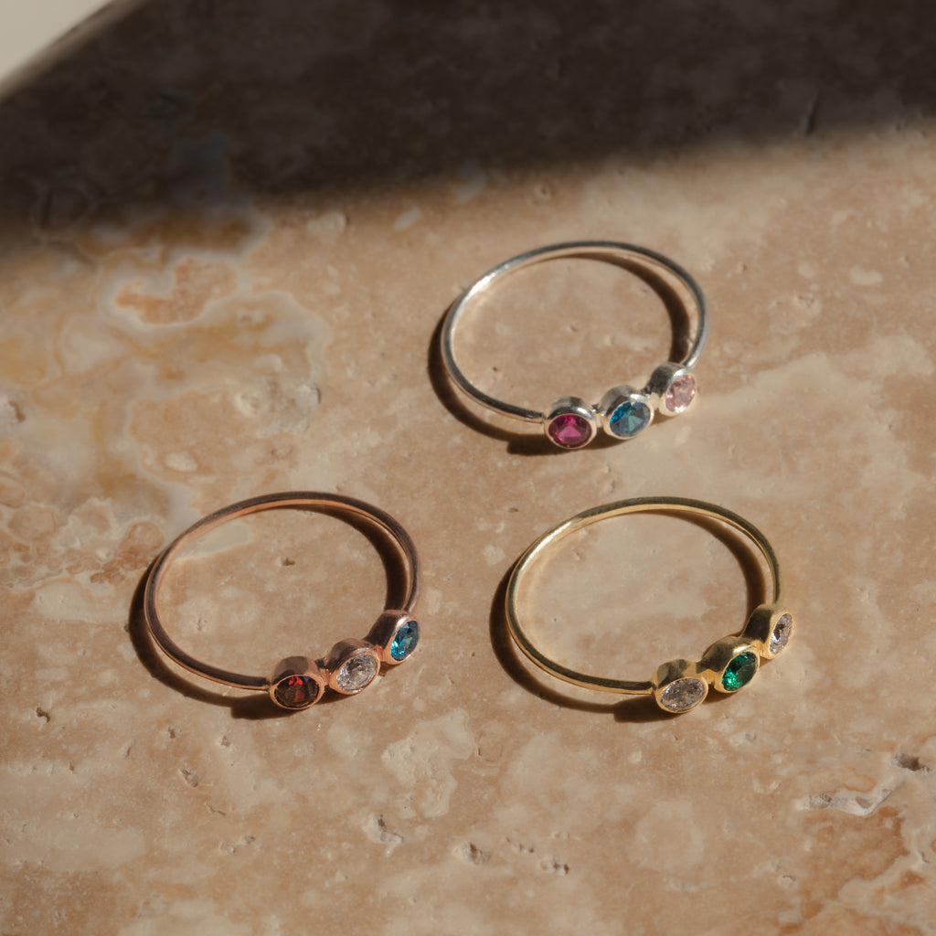The Triple Birthstone Ring, featuring three customizable colorful gemstones, rests on a marbled surface and glows in natural light.