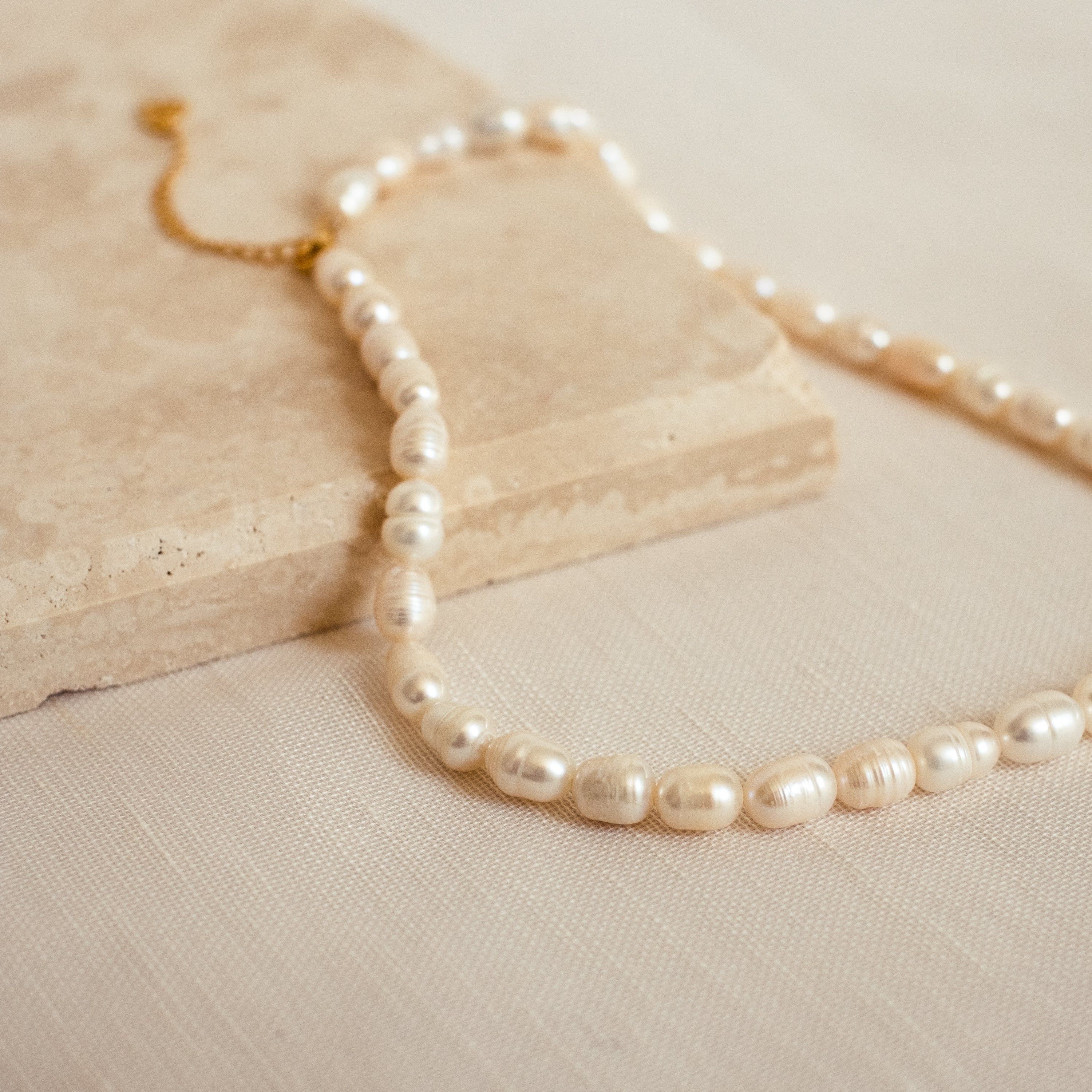 The Bold Pearl Necklace with a gold clasp is displayed on a textured beige stone and fabric surface.