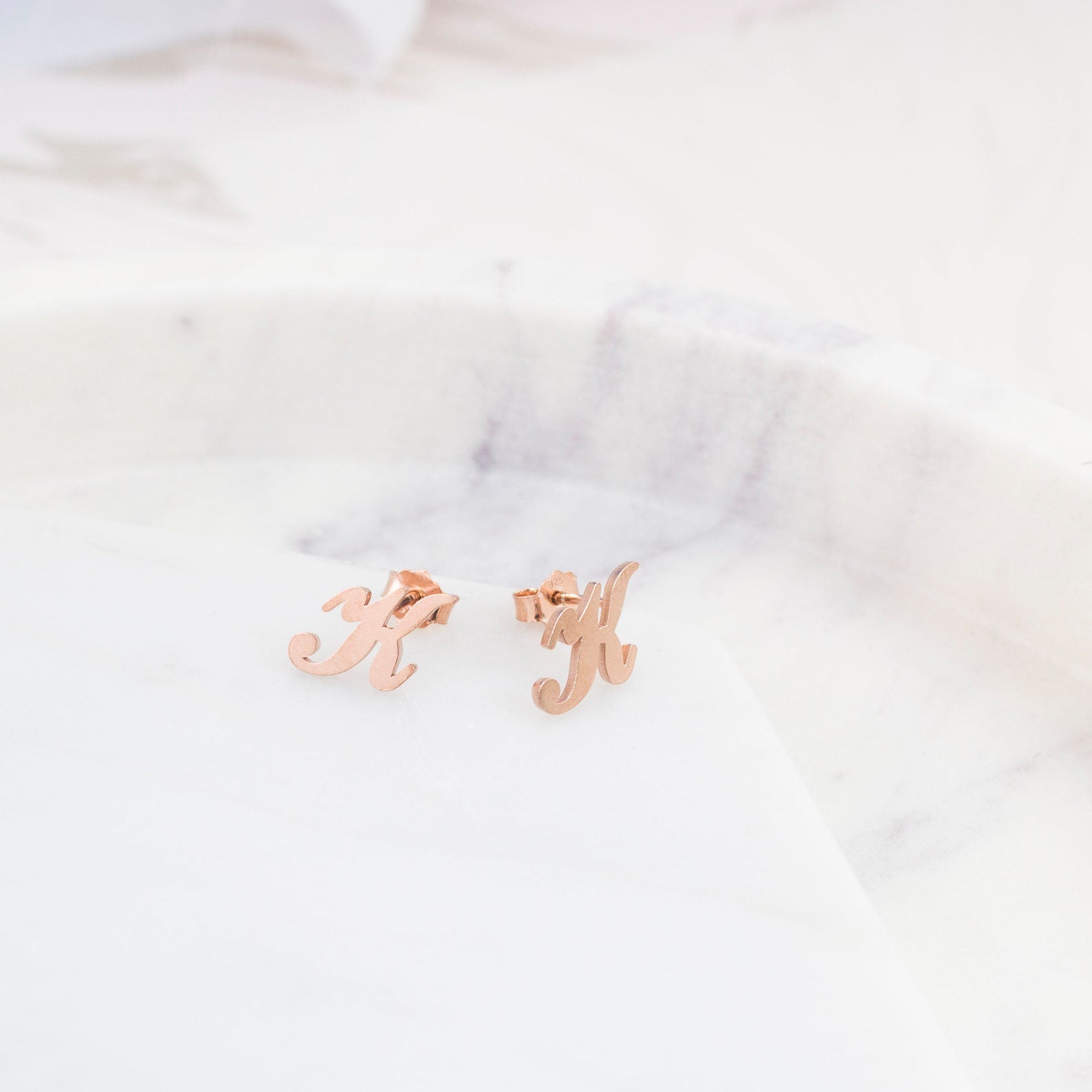 Alex Initial Studs in rose gold, shaped as the letter H, rest elegantly on a white marble surface—perfect personalized earrings for any occasion.
