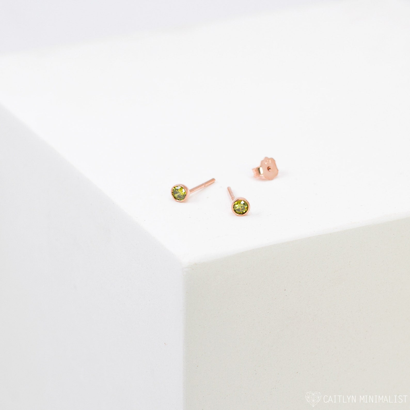 Birthstone Studs in rose gold with small green gemstones are displayed on a white geometric surface—ideal everyday earrings for a personalized style.
