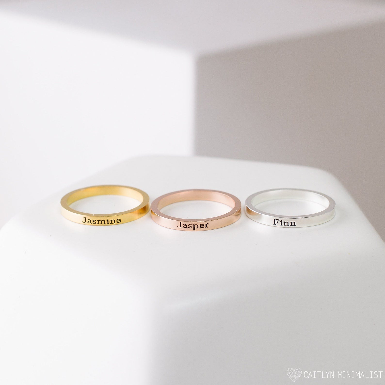 The Stacking Name Band features three custom engraved rings in gold, rose gold, and silver—shown with Jasmine, Jasper, and Finn—on a white surface. Ideal personalized jewelry for any meaningful occasion.