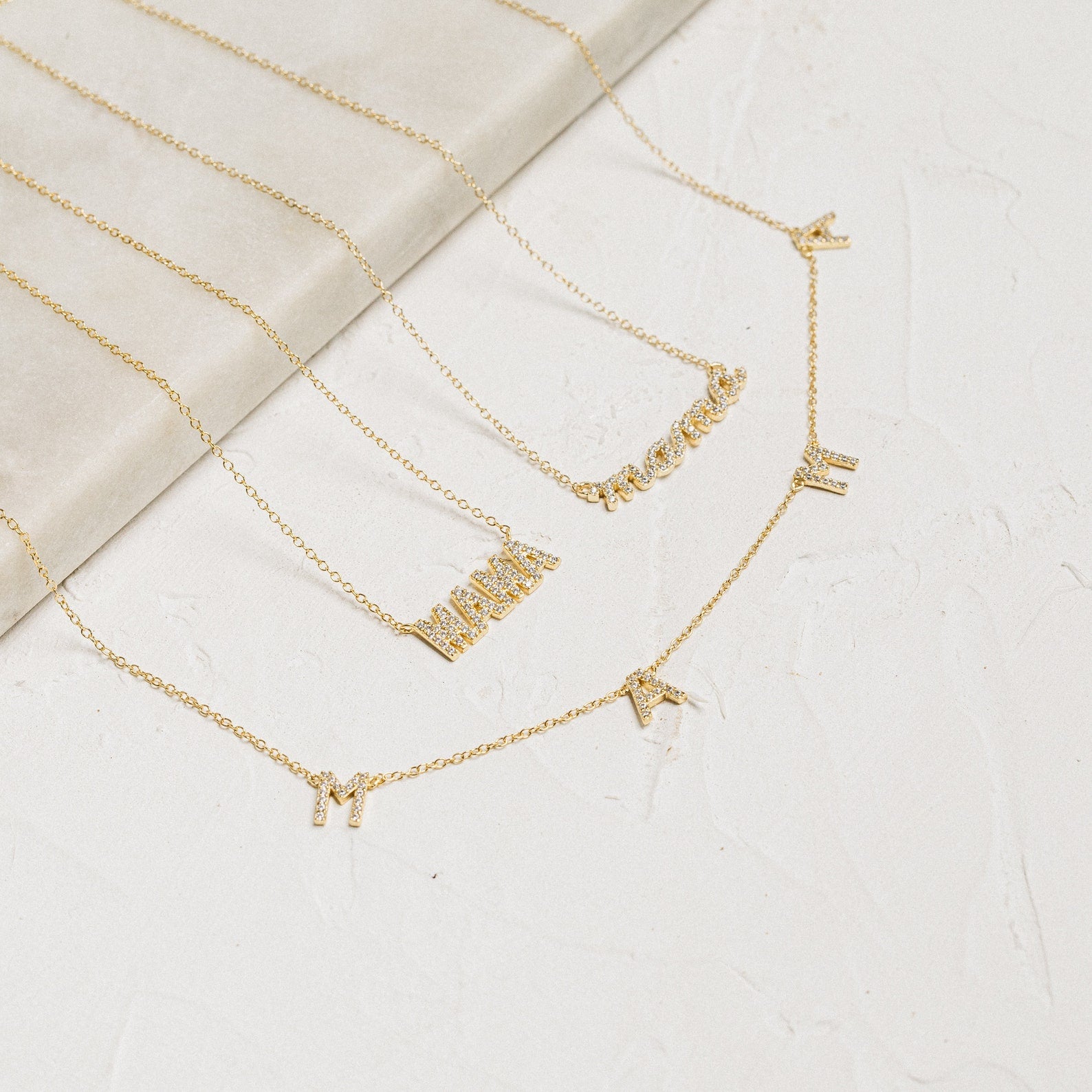 Three gold necklaces with letter charms are shown on a light textured surface; one is a stylish Mama Necklace—perfect for gifting—while the others spell MAVERICK and feature individual letters M, A, R, and I.