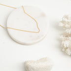 A gold curb chain necklace resting in a gentle curve on a round white marble tray with soft white coral in the background.