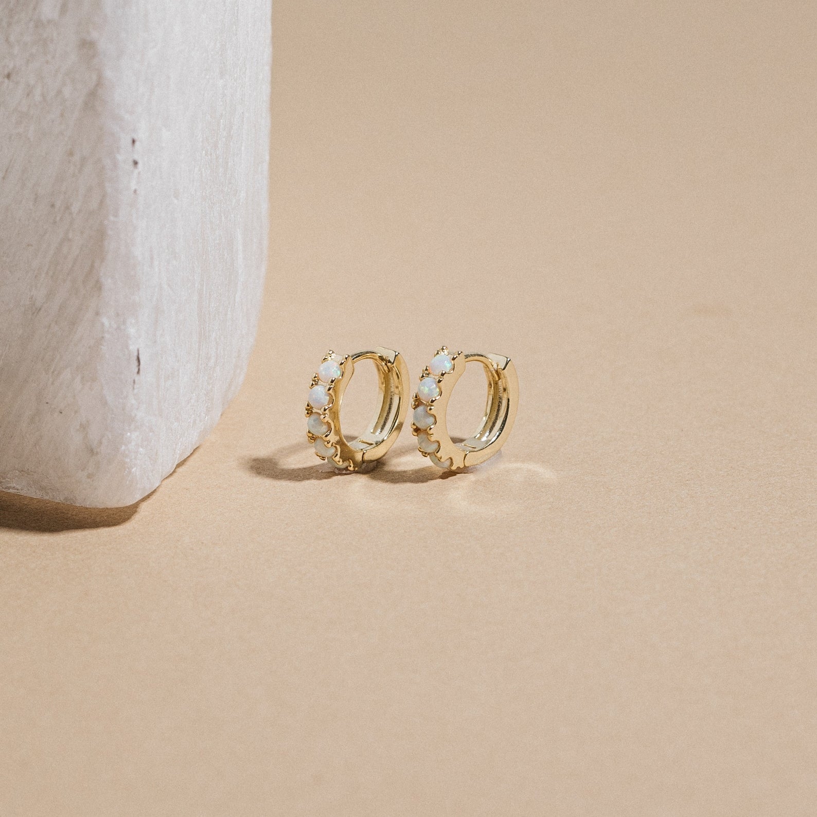 A pair of Opal Huggies, minimal gold earrings, displayed next to a white stone.