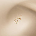 Elle Huggies—gold midi hoops with small rectangular pendants, inspired by the Elle Baguette Hoops—displayed on a smooth beige surface.