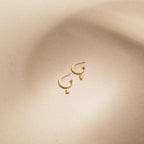 Elle Huggies—gold midi hoops with small rectangular pendants, inspired by the Elle Baguette Hoops—displayed on a smooth beige surface.