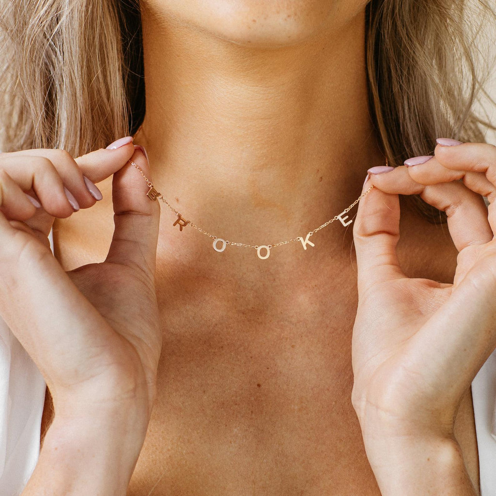 A woman displays a gold Custom Letter Necklace with charms spelling BROOKE by her collarbone, highlighting personalized, stylish jewelry.