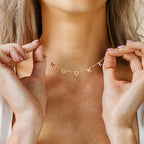 A woman displays a gold Custom Letter Necklace with charms spelling BROOKE by her collarbone, highlighting personalized, stylish jewelry.