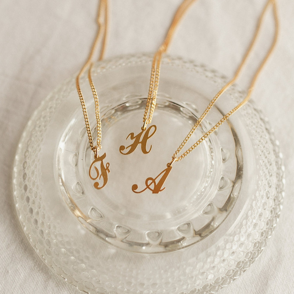Three gold Old English Initial Necklaces with Curb Chains—featuring the letters F, H, and A—are displayed on a clear glass dish for a chic goth-glam look.