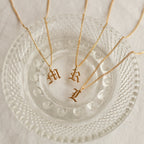Three gold Old English Initial Necklace with Curb Chain pieces—featuring “M,” “R,” and “L” pendants—rest elegantly on a clear glass dish.
