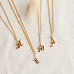 Four Old English Initial Necklaces with Curb Chain, featuring H, L, M, and A letter pendants in gold, are displayed on a light textured fabric for a stylish goth-glam look.