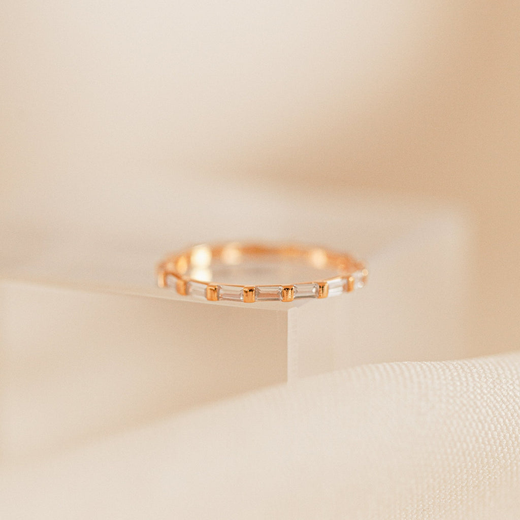 The Baguette Eternity Ring, featuring small rectangular gems, sparkles atop a clear acrylic block, capturing modern luxury against a soft beige background.