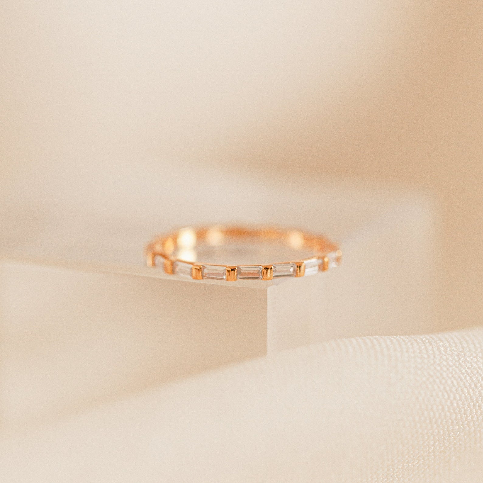 The Baguette Eternity Ring, featuring small rectangular gems, sparkles atop a clear acrylic block, capturing modern luxury against a soft beige background.