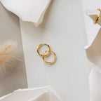 Two Kaia Diamond Huggies rest on a white surface, accented by abstract white shapes and nearby beige decorations.