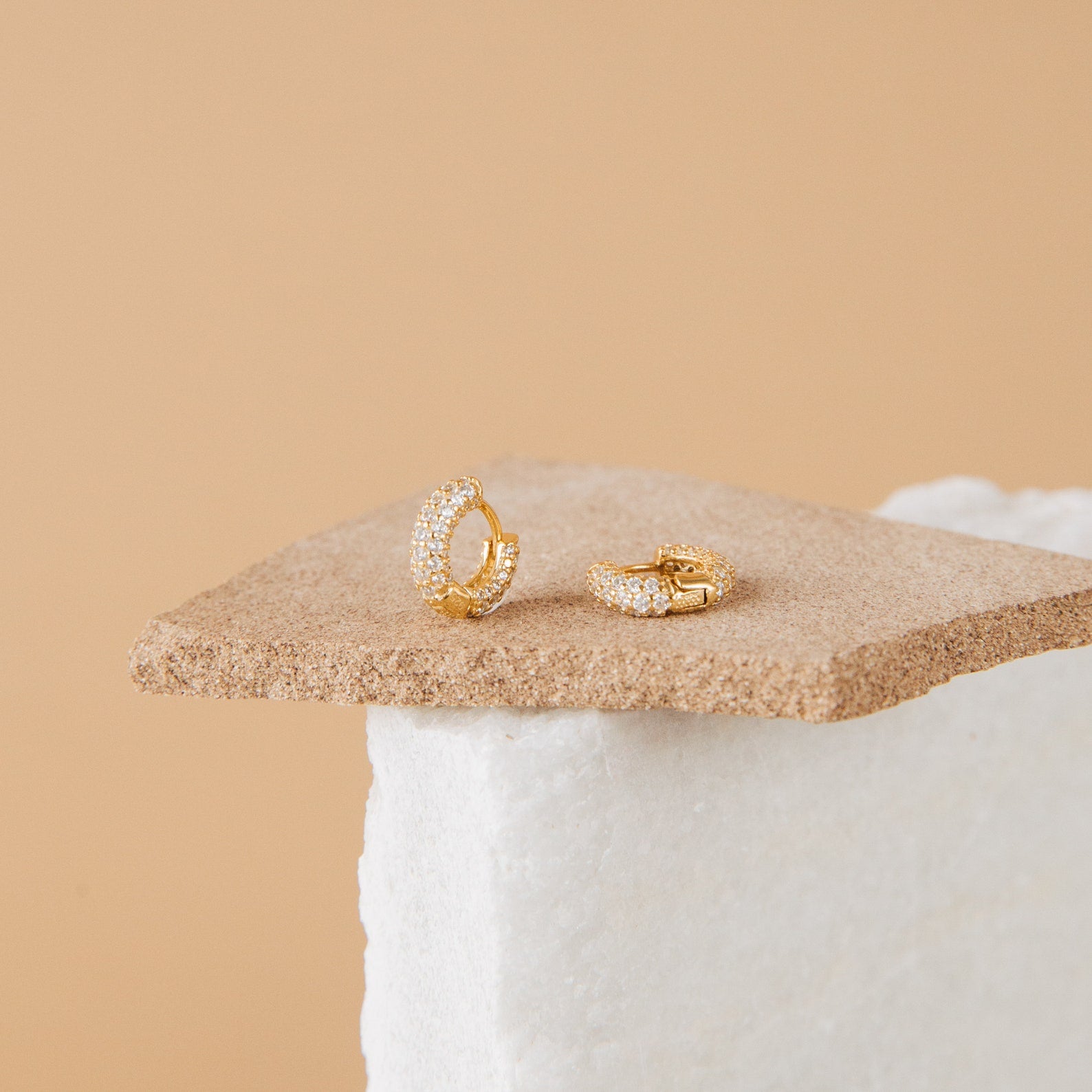 A pair of Kira Pave Huggies in small gold with pavé stones, displayed on a textured beige surface.
