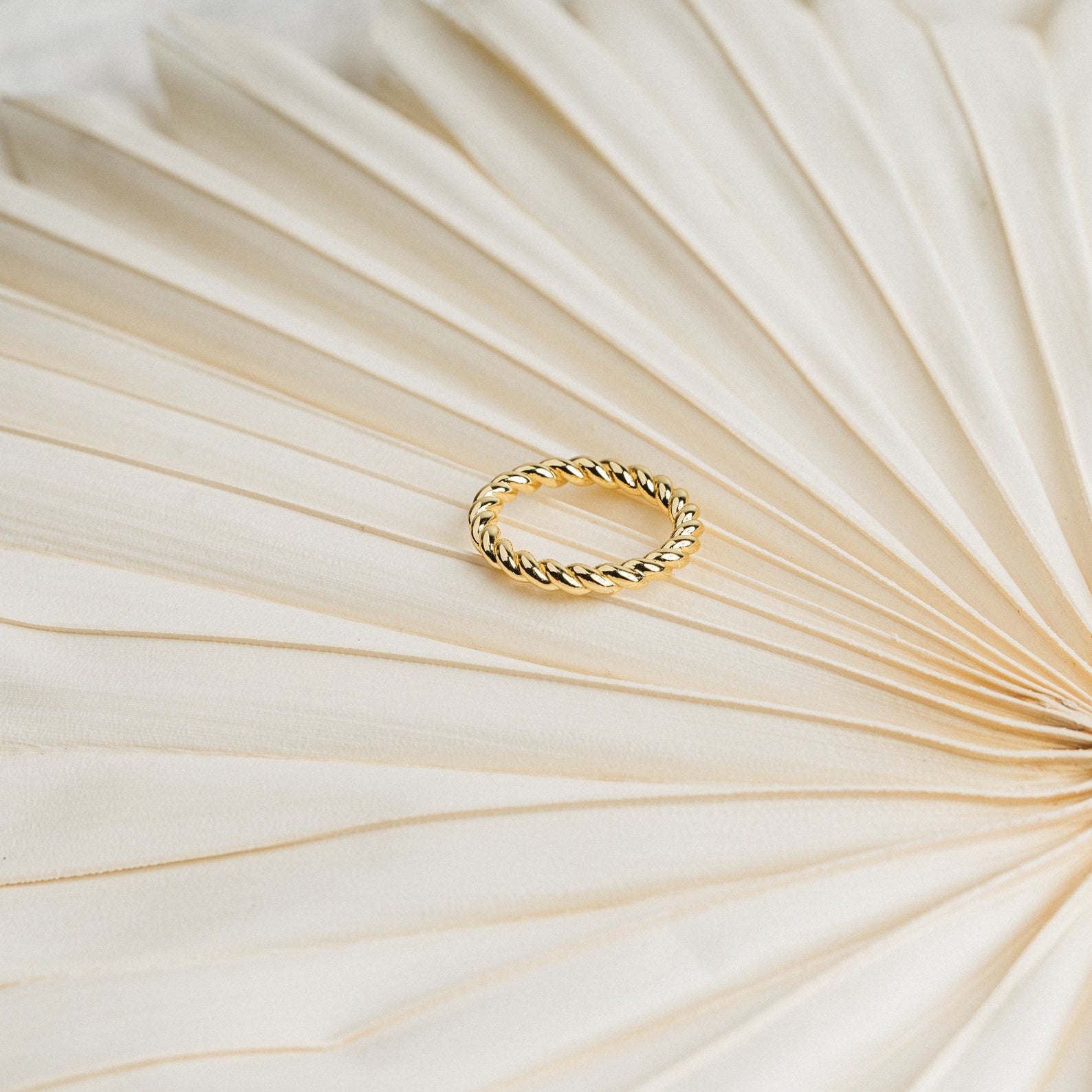 The Tia Twisted Ring in gold sits on a textured, cream fan-shaped surface—an elegant, modern piece ideal for stacking with your favorite rings.