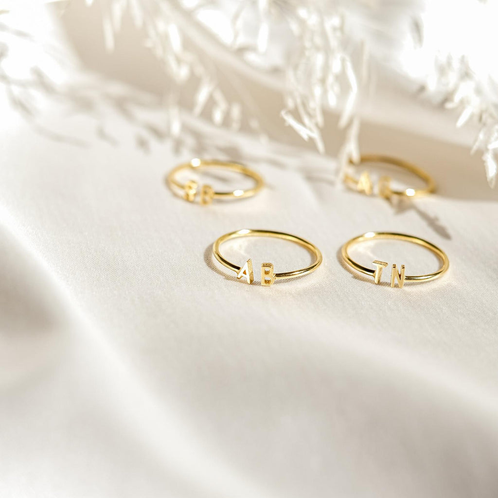 Four Modern Duo Initial Rings (AB, IN, etc.) shine on a soft white surface in sunlight—ideal custom jewelry to personalize your ring collection.