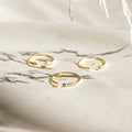 Three gold Venice Initial Birthstone Rings rest on light fabric, each with a delicate gemstone and cursive lowercase letter charm—a personalized ring, perfect as an initial birthstone accessory for everyday elegance.