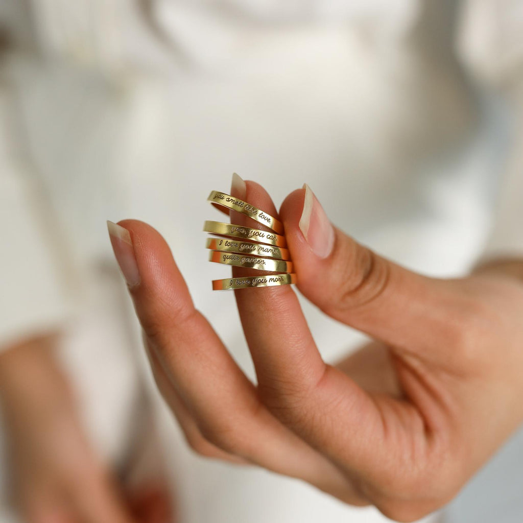 A hand holds four Ann Thin Message Rings in Rose Gold, each engraved with "I love you more," against a soft, neutral background—an ideal holiday gift.
