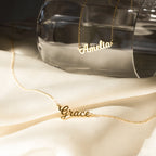 Two rose gold Mellow Name Necklaces, personalized with the names Amelia and Grace, are displayed on cream fabric and a glass surface.