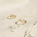 Two delicate Venice Name Rings with elegant cursive script rest on a textured cream surface, accented by dried plant stems.