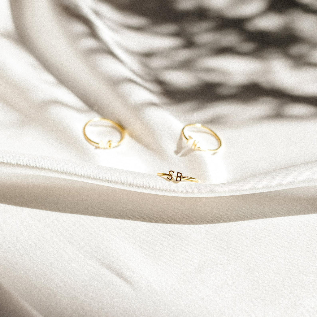 Three gold rings rest on soft white fabric: one Modern Duo Initial Ring engraved with S B and two plain, all touched by dappled light and shadow.