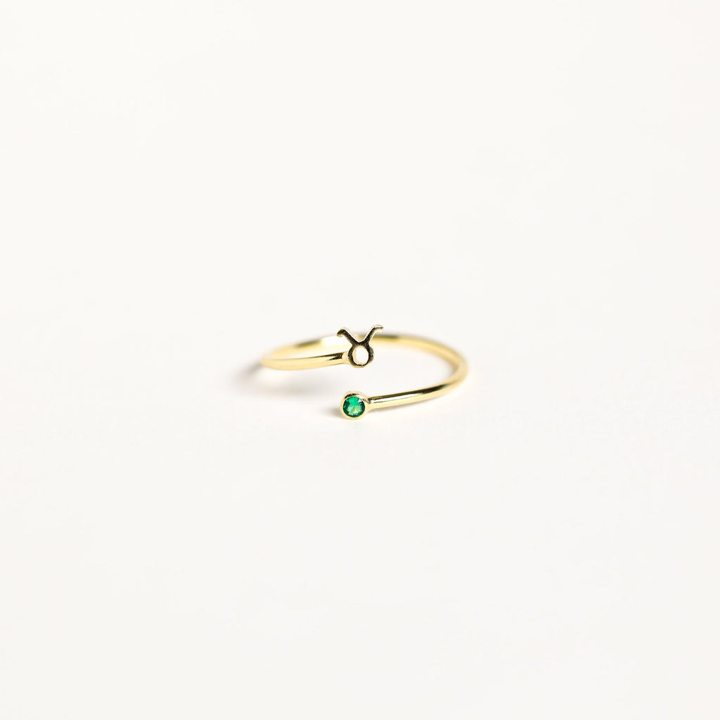 The Zodiac Birthstone Ring features a gold band with the Taurus symbol and a small green birthstone, combining elegant jewelry with celestial charm on a plain white background.