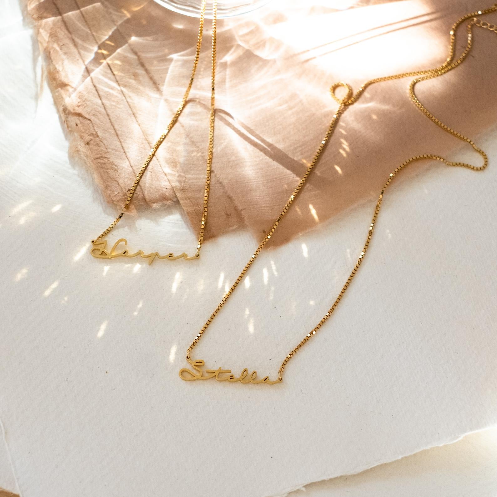 Two gold Summer Name Necklaces in Box Chain reading Harper and Stella rest on a sunlit, textured surface—an elegant addition to your jewelry collection.