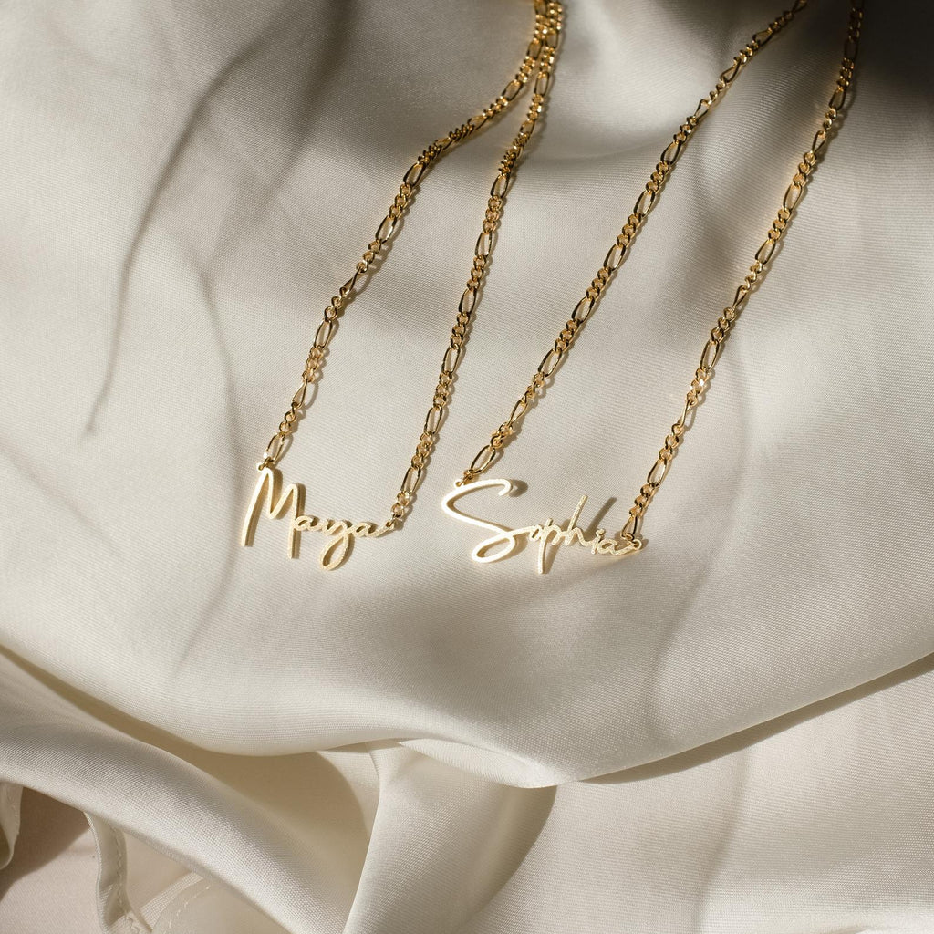 Two Summer Name Necklaces in Figaro Chain, with gold cursive nameplates reading Maya and Sophia, are displayed on a cream-colored silky fabric background.