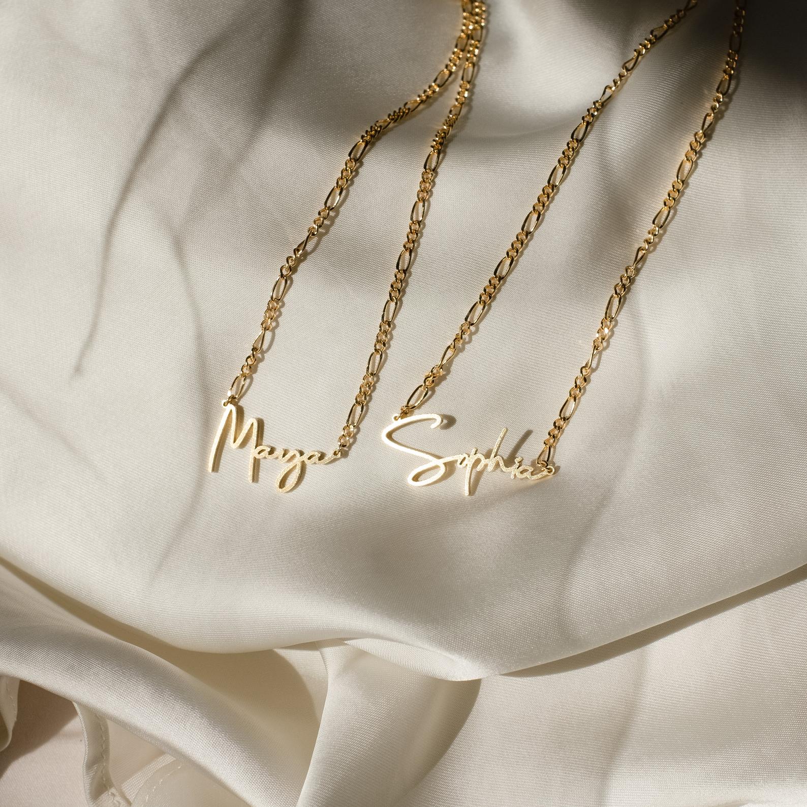 Two Summer Name Necklaces in Figaro Chain, with gold cursive nameplates reading Maya and Sophia, are displayed on a cream-colored silky fabric background.