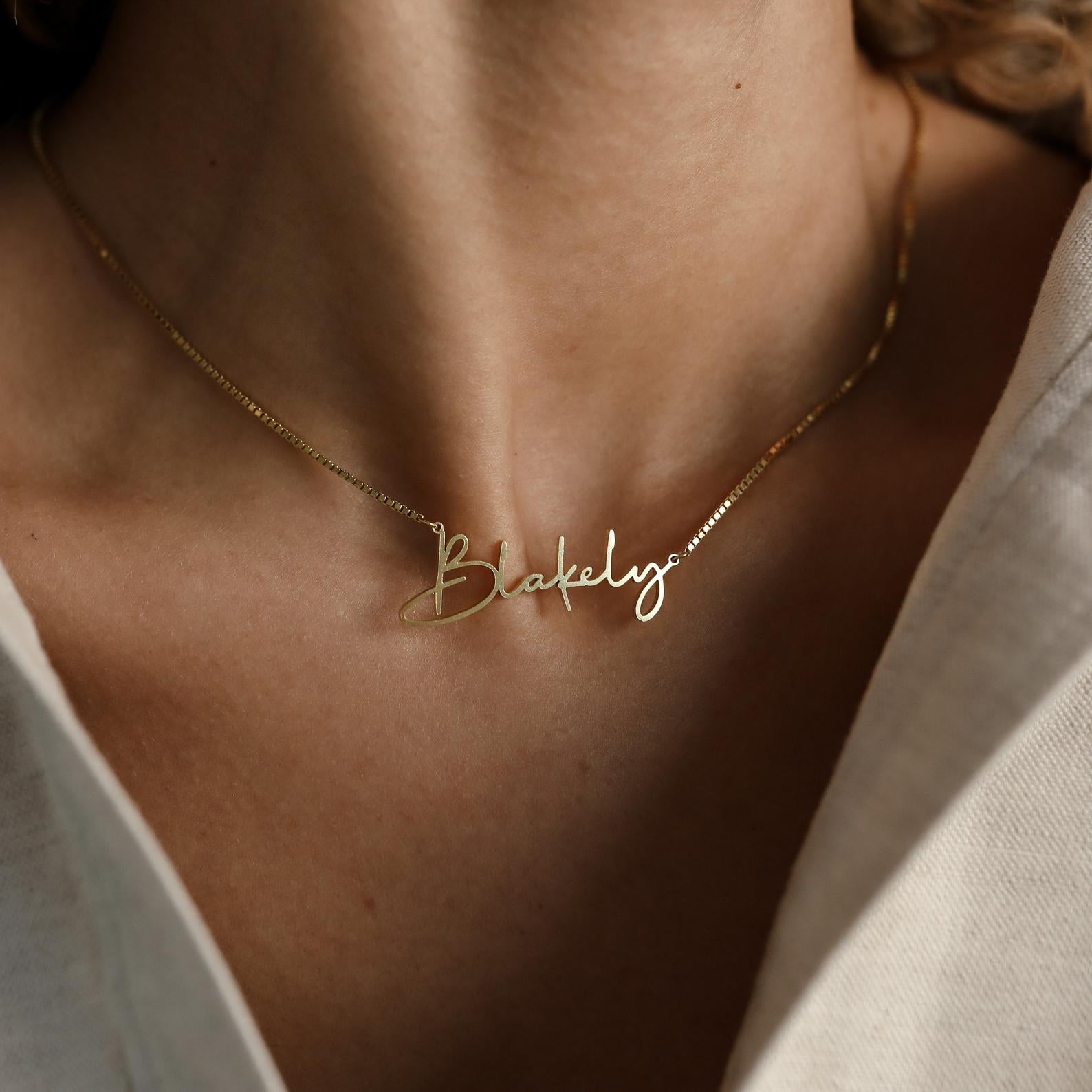The Summer Name Necklace in Box Chain features a cursive Blakely pendant, elegantly resting on the neck and paired with a light-colored top—a versatile accessory for any occasion.
