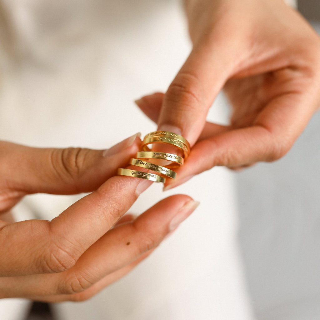 A person showcases four Ann Thin Message Rings, each engraved with custom text, against manicured nails and soft lighting—a stylish personalized gift idea.