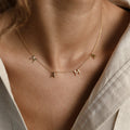 Gold “MAMA” letter necklace worn at the collarbone, featuring four spaced capital letter charms on a delicate chain, styled against a neutral-toned outfit.