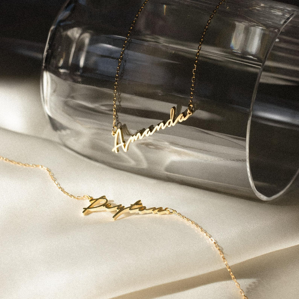 Two gold Minimal Name Necklaces, one with "Amanda" on a glass and another with "Peyton" on cream fabric, make the perfect personalized gift.