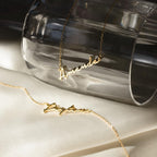 Two gold Minimal Name Necklaces, one with "Amanda" on a glass and another with "Peyton" on cream fabric, make the perfect personalized gift.