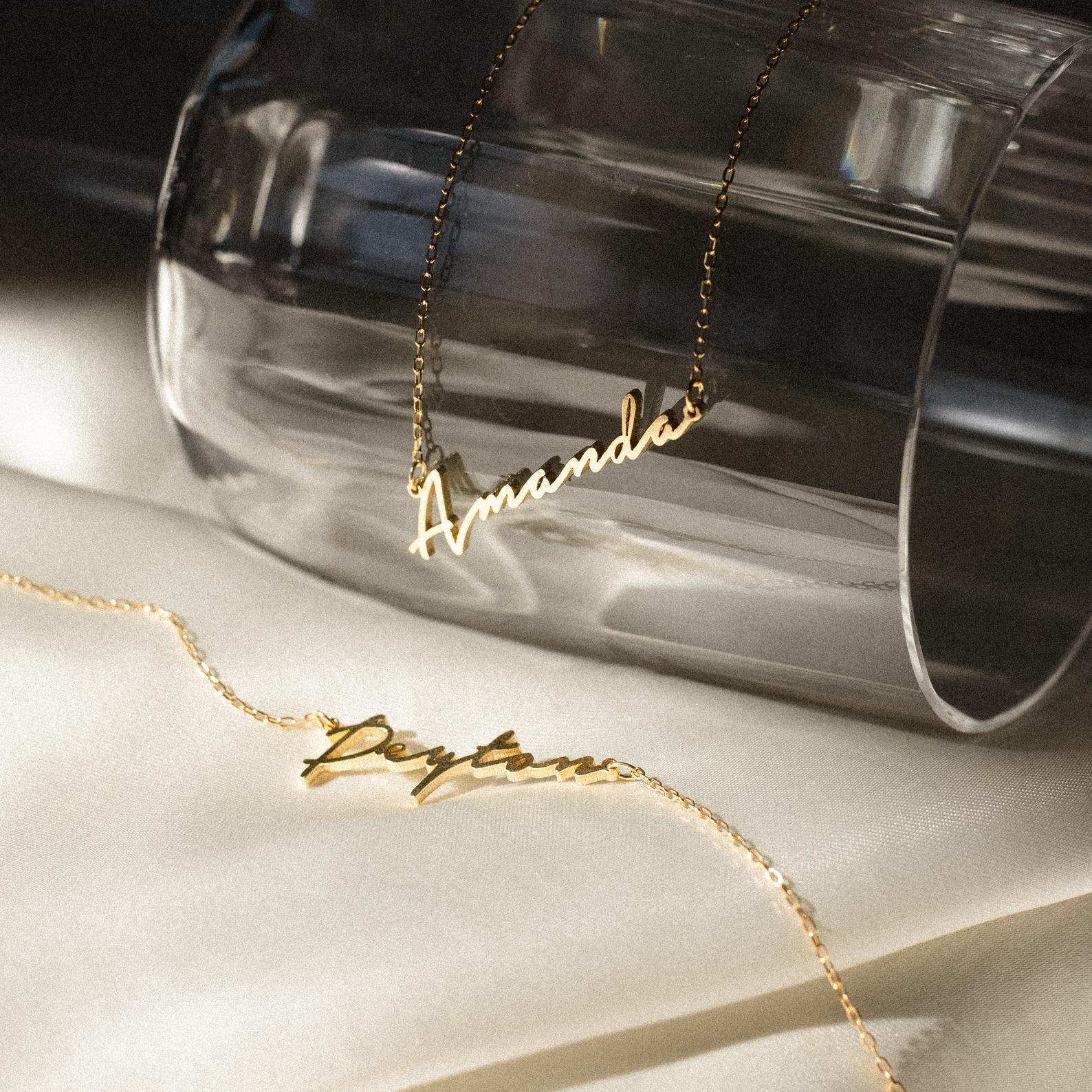 Two gold Minimal Name Necklaces, one with "Amanda" on a glass and another with "Peyton" on cream fabric, make the perfect personalized gift.