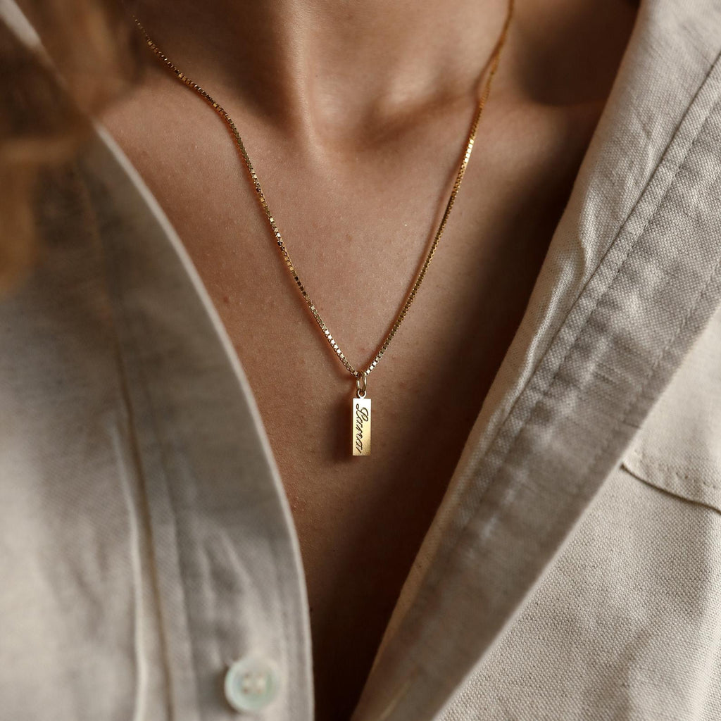 Dainty Tag Name Necklace featuring a rectangular engraved pendant styled on delicate chains, paired with a beige button-up shirt.