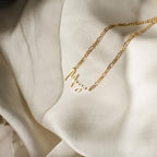 The Carrie Name Necklace in Figaro Chain, styled in gold with "Maya," rests elegantly on soft cream-colored fabric, showcasing its minimal design.