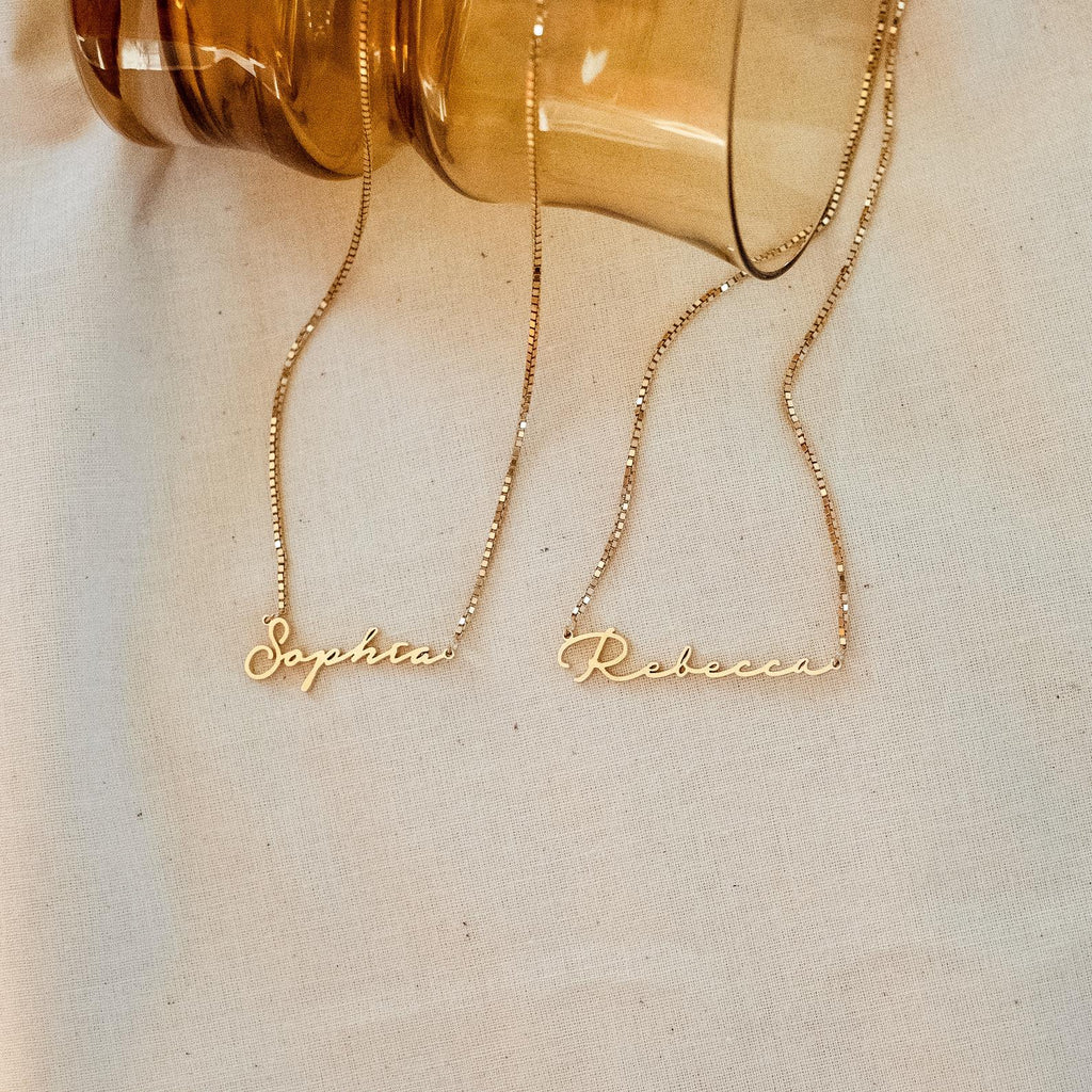 Two dainty necklaces reading Sophia and Rebecca rest on cream fabric near a brown glass jar, each featuring a delicate box chain just like the elegant Venice Name Necklace in Box Chain.