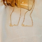 Two dainty necklaces reading Sophia and Rebecca rest on cream fabric near a brown glass jar, each featuring a delicate box chain just like the elegant Venice Name Necklace in Box Chain.