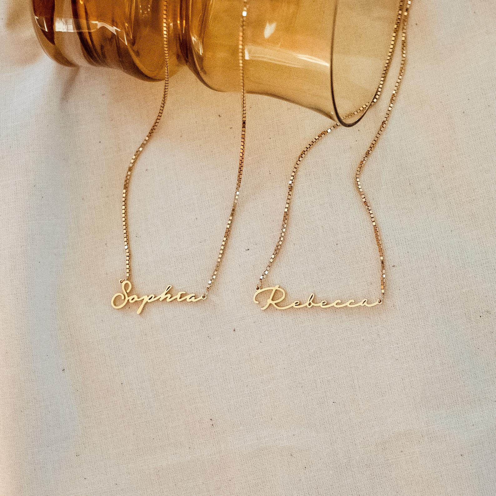 Two dainty necklaces reading Sophia and Rebecca rest on cream fabric near a brown glass jar, each featuring a delicate box chain just like the elegant Venice Name Necklace in Box Chain.