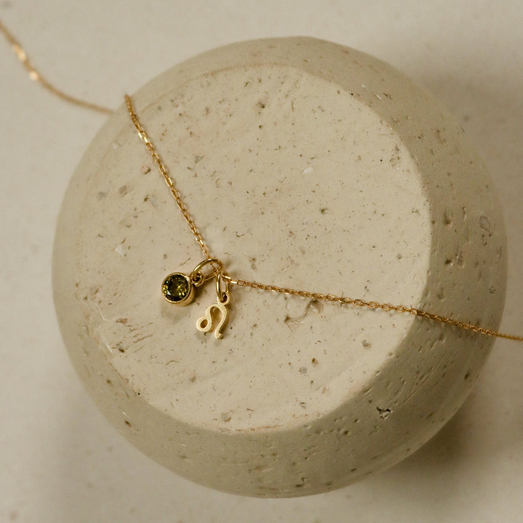 The Zodiac Birthstone Necklace features a gold chain with a small green birthstone and Leo zodiac charm, elegantly displayed on a round stone surface—perfect for personalized style.
