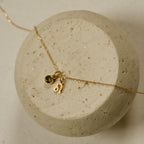 The Zodiac Birthstone Necklace features a gold chain with a small green birthstone and Leo zodiac charm, elegantly displayed on a round stone surface—perfect for personalized style.