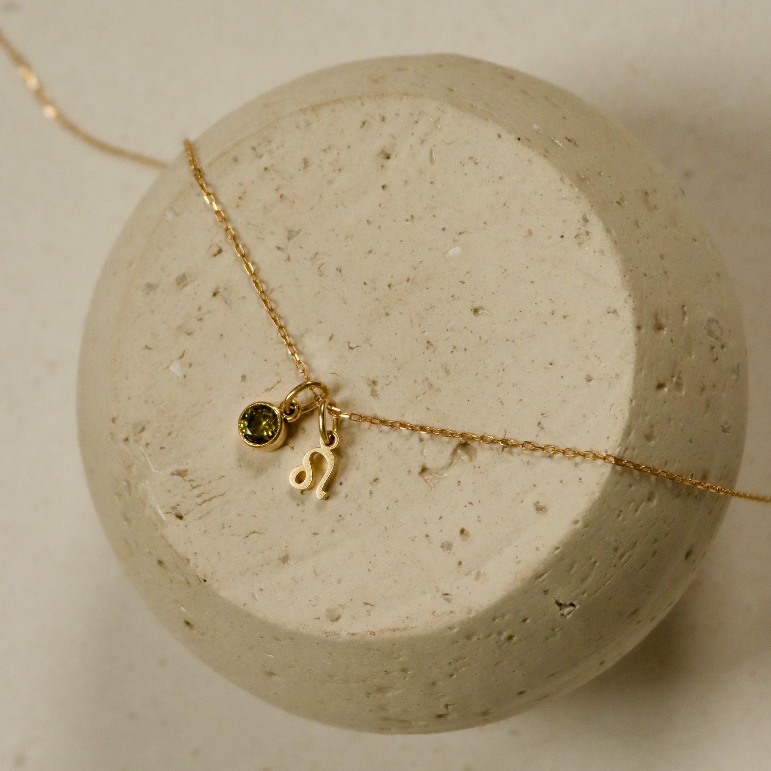 The Zodiac Birthstone Necklace features a gold chain with a small green birthstone and Leo zodiac charm, elegantly displayed on a round stone surface—perfect for personalized style.