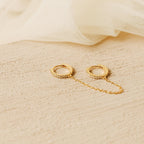 Two Pave Duo Chain Earrings, ideal for multiple ear piercings, are linked by a delicate chain and displayed on a textured beige surface with soft fabric in the background.