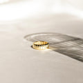 A gold Audrey Ring with a grooved pattern rests on a sunlit surface, casting a gentle shadow—an essential piece ideal for everyday wear.