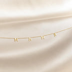 The Mama Letter Necklace, with charms spelling MAMA, is displayed on soft cream-colored fabric—a perfect gift to celebrate motherhood.