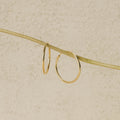 The Endless Thin Hoops, displayed on a slender pale branch with a textured beige background, are timeless gold earrings crafted to be the perfect everyday accessory.