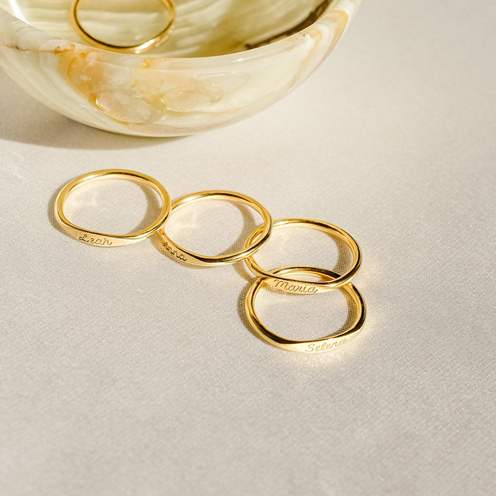 The Ann Skinny Name Ring in Rose Gold, engraved with names, is shown near a marble bowl on a light textured surface—making it a perfect Christmas gift for her.